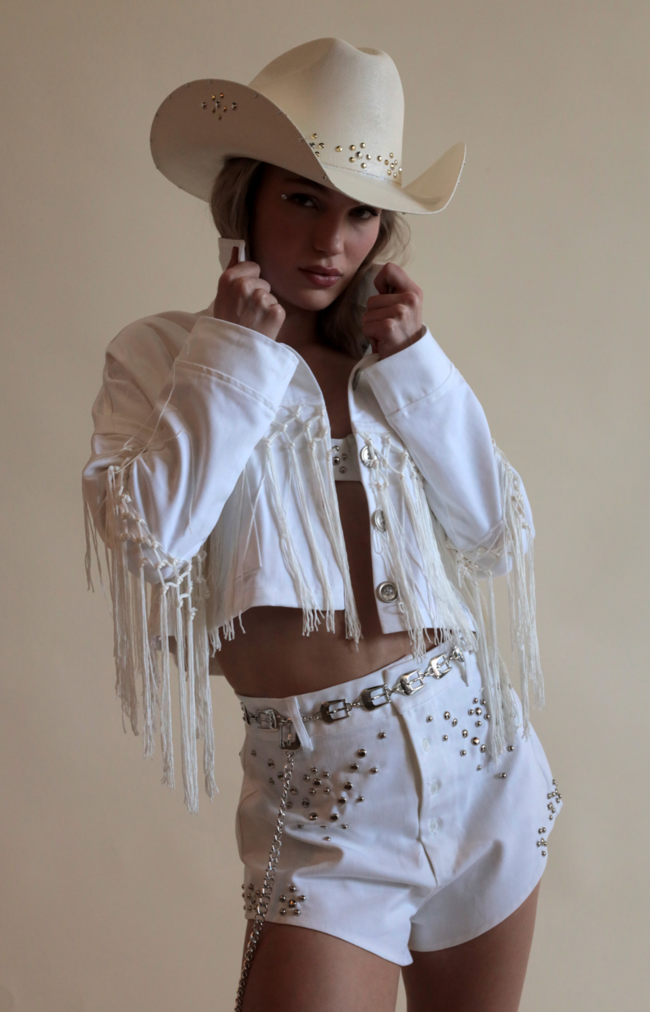 White cowboy hat with metallic jewels paired with denim grommeted set and fringe jacket.
