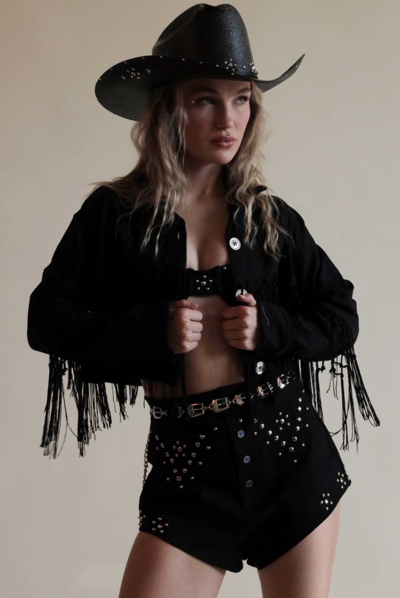Black cowboy hat with metallic jewels paired with denim grommeted set and fringe jacket.